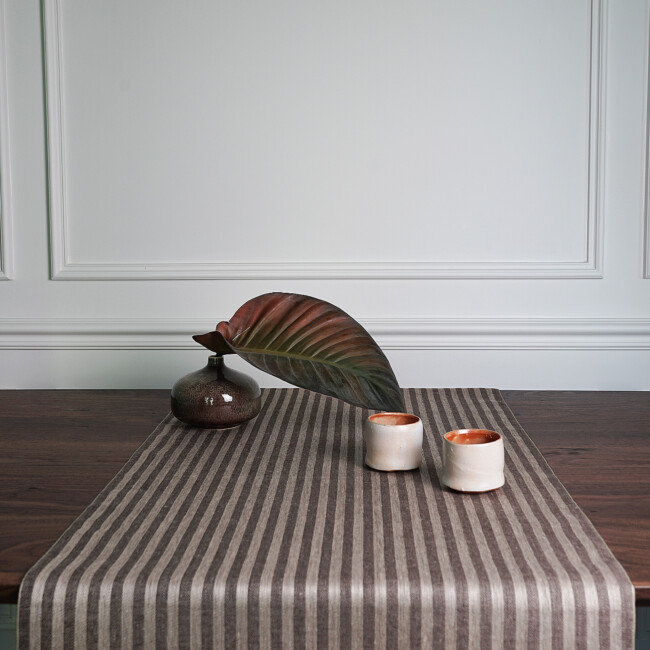 The striped Anne table runner in chocolate brown/sand on a dark brown table, by Klassbols Linnevaveri