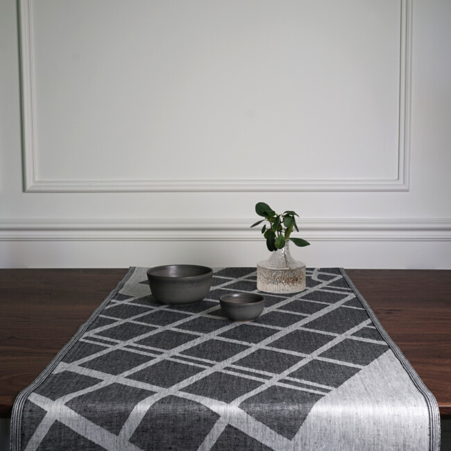 The table runner Arvika in the color black, lies on a walnut-colored table, with a detail of a vase and green wax, by Klassbol&#39;s Linnevaveri