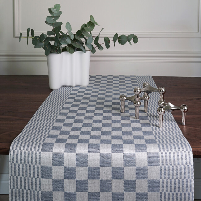 Schackrutan table runner in denim blue on a dark brown dining table in front, with a white vase that has green leaf sticks in it, from Klassbol&#39;s linen weave