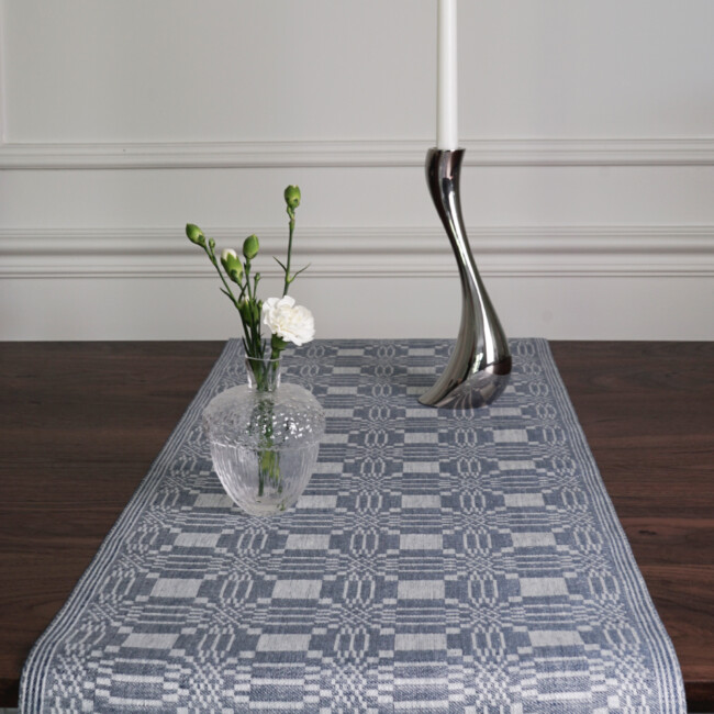 Lemon table runner in the color denim blue on a walnut colored table, together with a dark lamp and a candle lantern, from Klassbol&#39;s Linen Vaveri